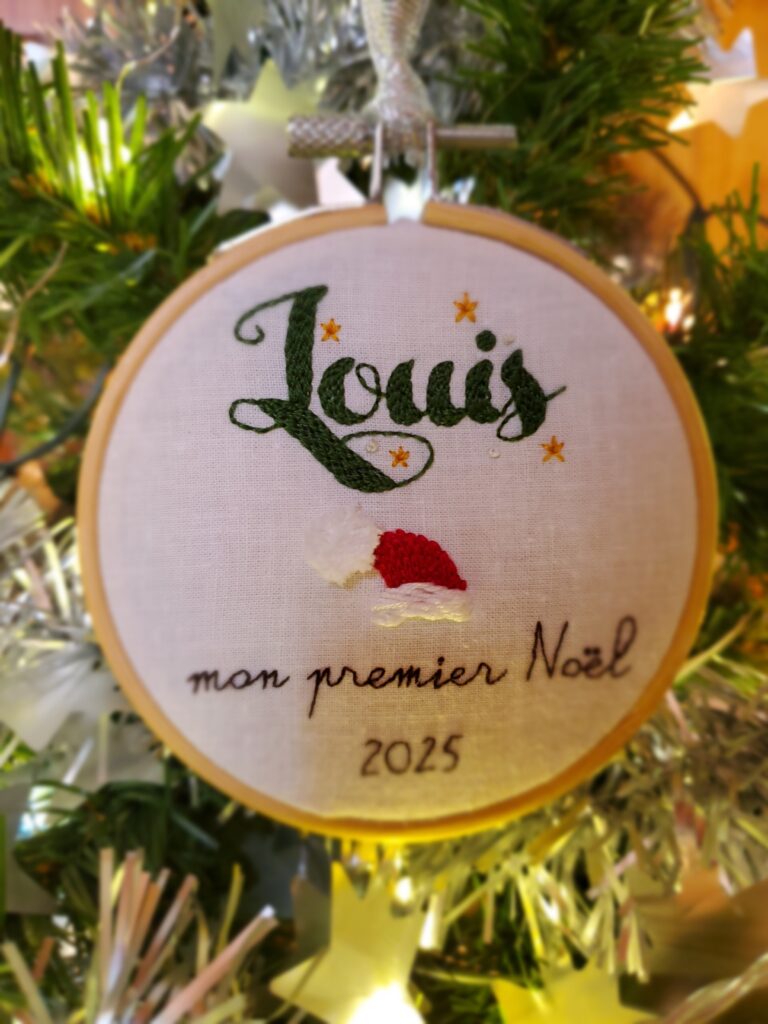 Boule de Noël brodée à la main comportant le prénom du bébé et un bonnet de lutin + la mention "mon premier Noël" et la date 2025